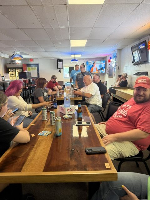 People sit around a wooden bar table, chatting and drinking. Some use phones, while others talk. The scene is casual, with various cans and bottles, and a TV is on in the background.