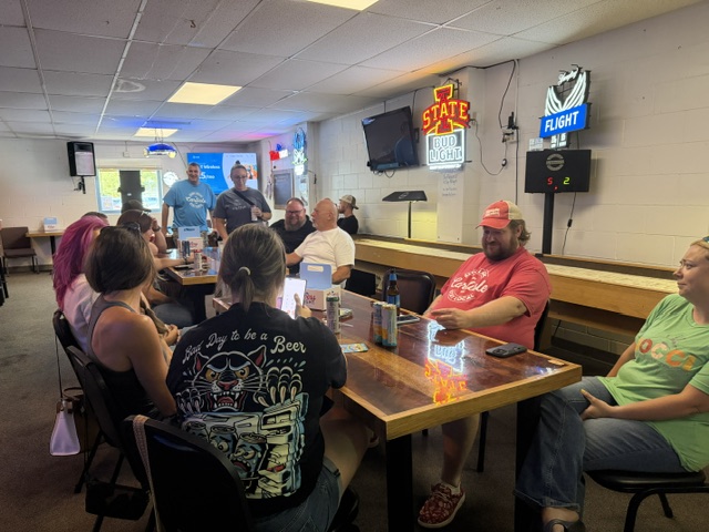 A group of people sit around a table in a casual bar, talking and smiling. Neon signs and drink menus are on the wall, and sunlight shines through a window in the background.