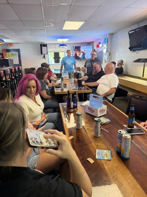 A group of people sit around a wooden table at a bar, chatting and playing a game. There are drinks, bingo cards, and cans on the table. The atmosphere appears relaxed and casual.
