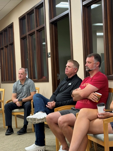 Four adults sit on chairs indoors, attentively listening. Three men are visible; one wears a gray shirt, one a black jacket, and one a maroon shirt holding a coffee cup. The setting appears to be a meeting room with wood-trimmed windows.