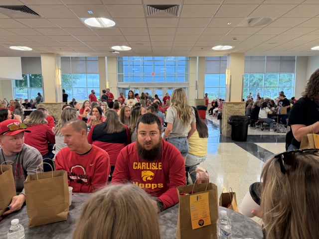 A group of people, many in red Carlisle Hoops shirts, sit at tables in a cafeteria with brown paper bags, talking and socializing. The room is bright with large windows in the background.
