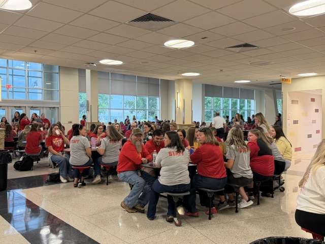 Large group of people, many wearing red or gray shirts, sit at round lunch tables in a bright cafeteria. Some shirts say “STAFF.” Windows line the back wall, and more people stand near the entrance.