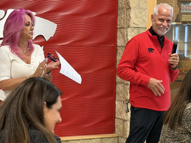 A man in a red shirt speaks into a microphone while a woman with pink hair holding papers stands beside him, clapping. Other people are seated in the foreground. Red and beige wall decor is visible in the background.