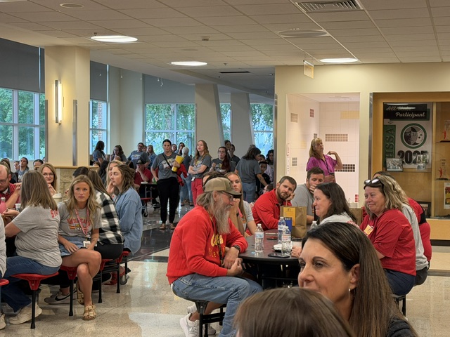 Large group of people sit and stand in a bright cafeteria, some at round tables, others mingling or walking. Most appear relaxed, wearing casual clothes. A display case and windows are visible in the background.