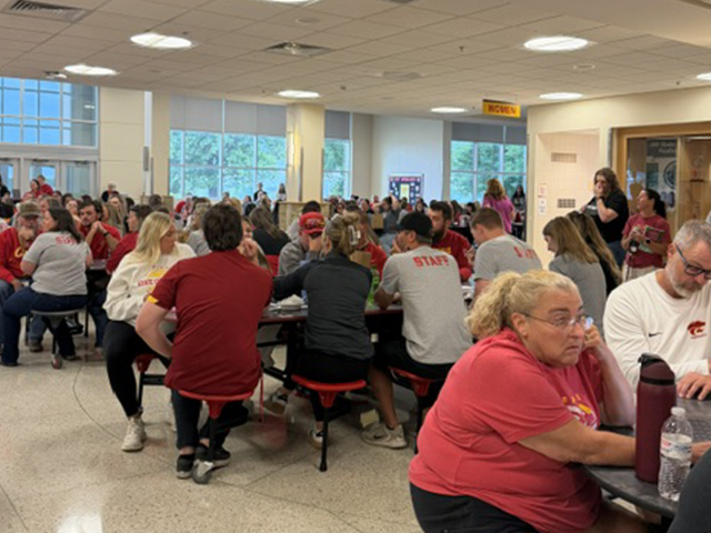 Large group of people, many wearing red or gray shirts, sit at round tables in a bright cafeteria. Some chat, while others work. Windows and a door are visible in the background.