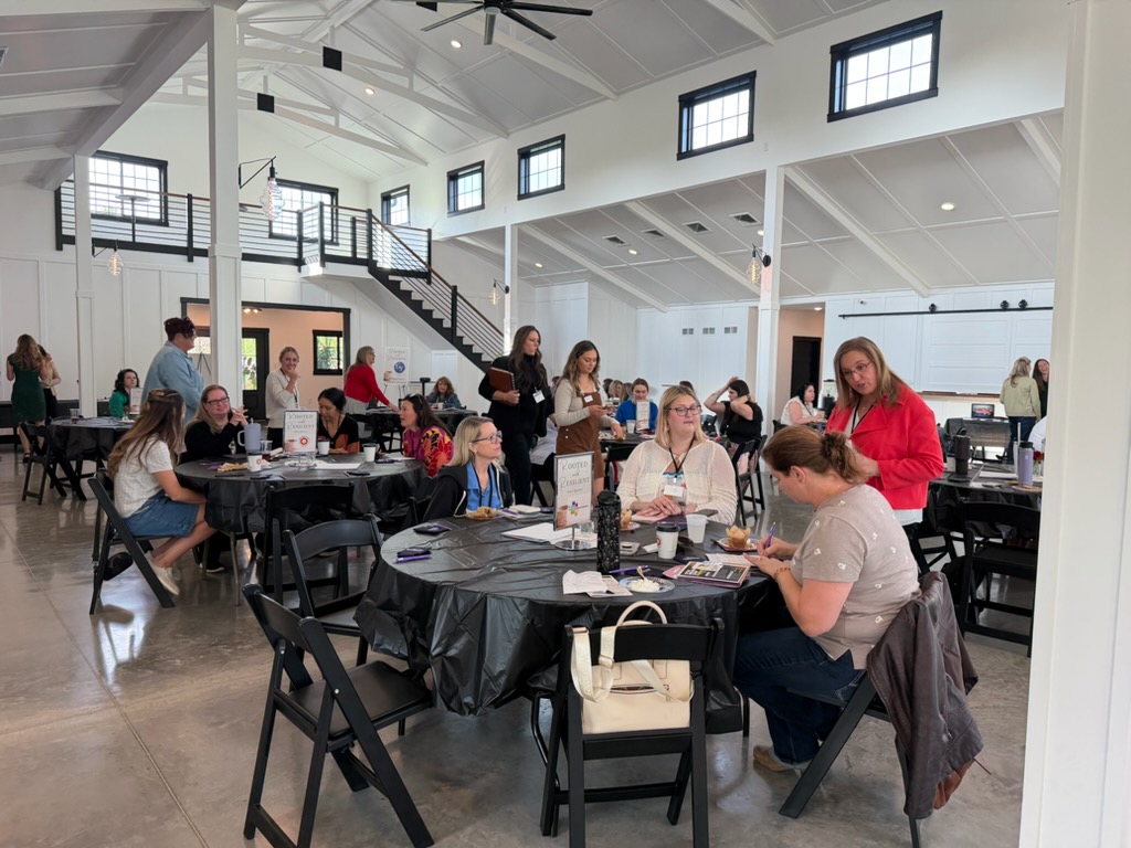 People sit at round tables in a bright, modern room with white walls and high ceilings. Some are talking, others work on papers. A few stand and interact. Large windows let in natural light.