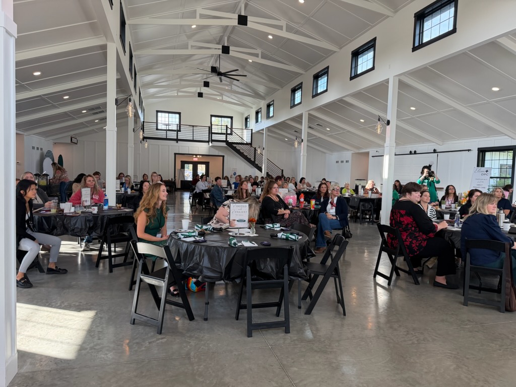 A large group of people sits at round tables in a bright, modern event space with high ceilings and tall windows, attentively listening to a presentation.
