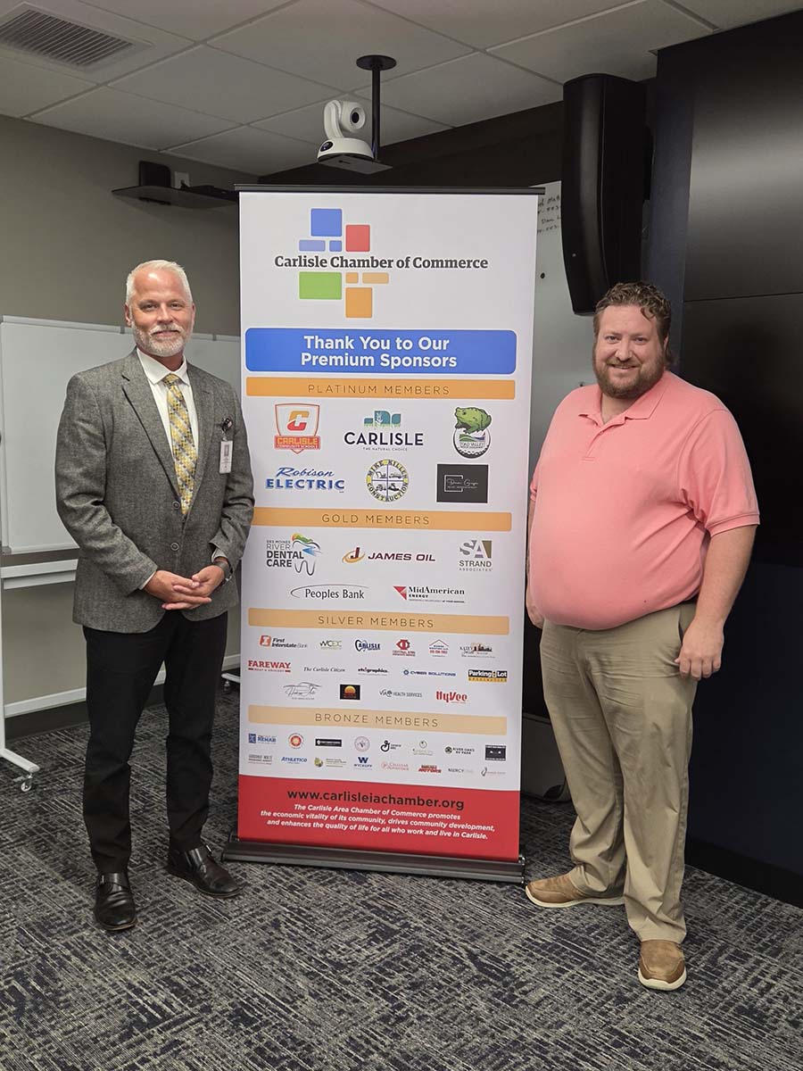 Two men stand beside a Carlisle Chamber of Commerce sponsor banner in an office. One wears a suit and tie, the other a pink polo and khakis. The banner displays logos and lists chamber sponsors by membership level.